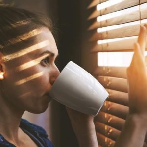 Woman,Looking,Through,Window,Blinds,Into,The,Sunlight,And,Drinking